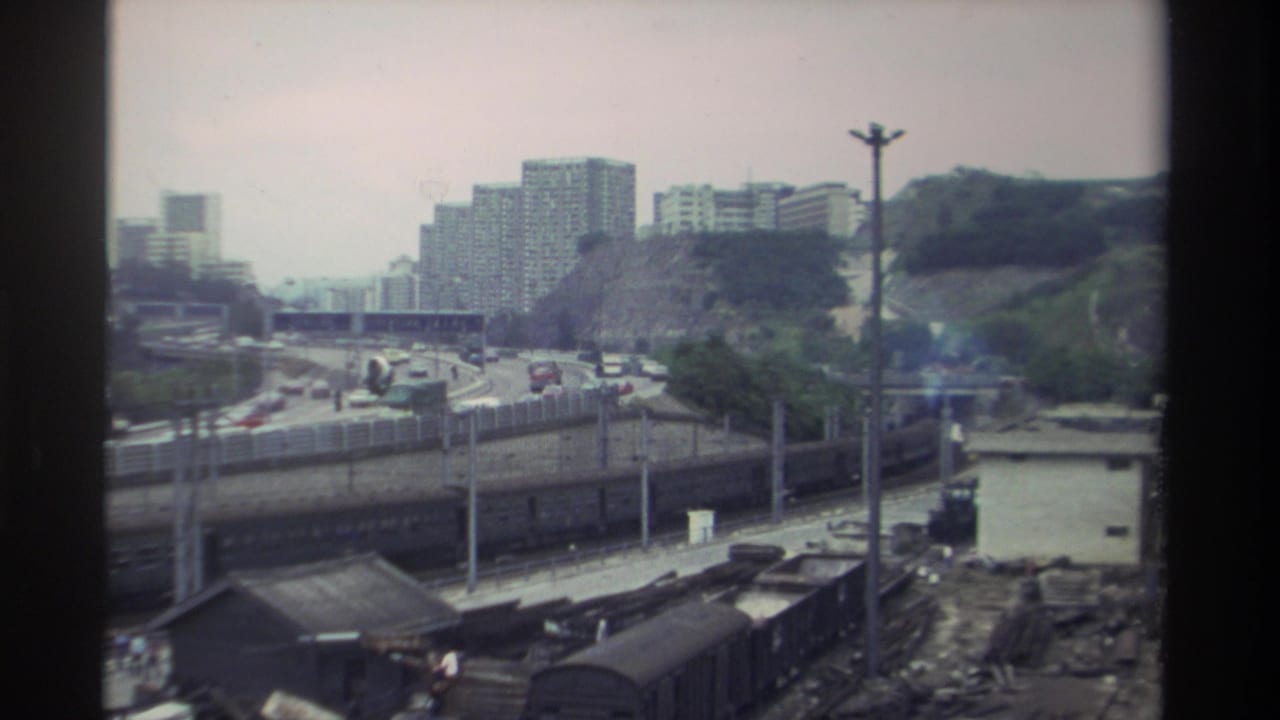 1982 Taiwan: 8mm window shot of rail freight infrastructure beside a congested highway, residential towers rising behind a tree-covered hillside.