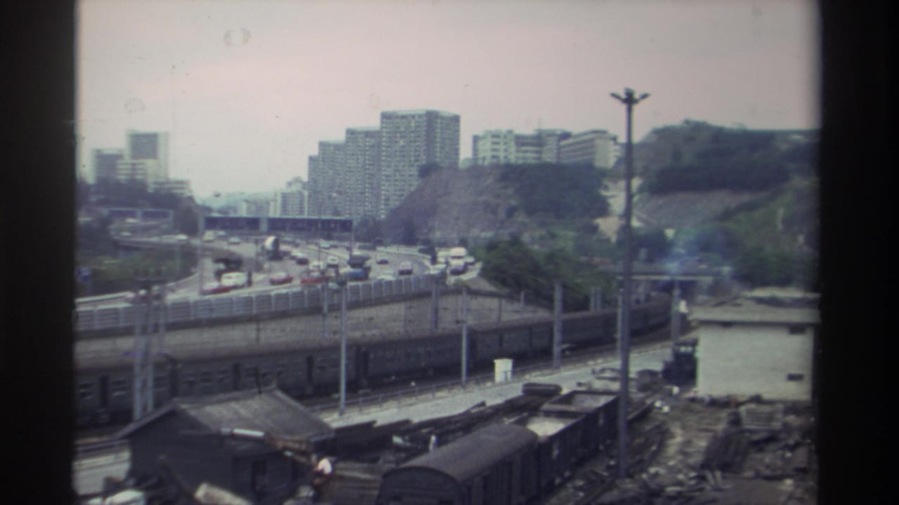 Elevated view of a freight rail yard in Taiwan, circa 1982, with dark freight wagons, a busy multi-lane highway, and mid-rise residential towers on a wooded hillside in the background.