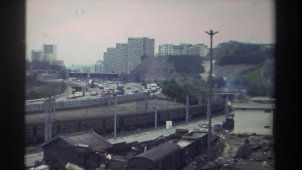 Elevated view of a freight rail yard in Taiwan, circa 1982, with dark freight wagons, a busy multi-lane highway, and mid-rise residential towers on a wooded hillside in the background.