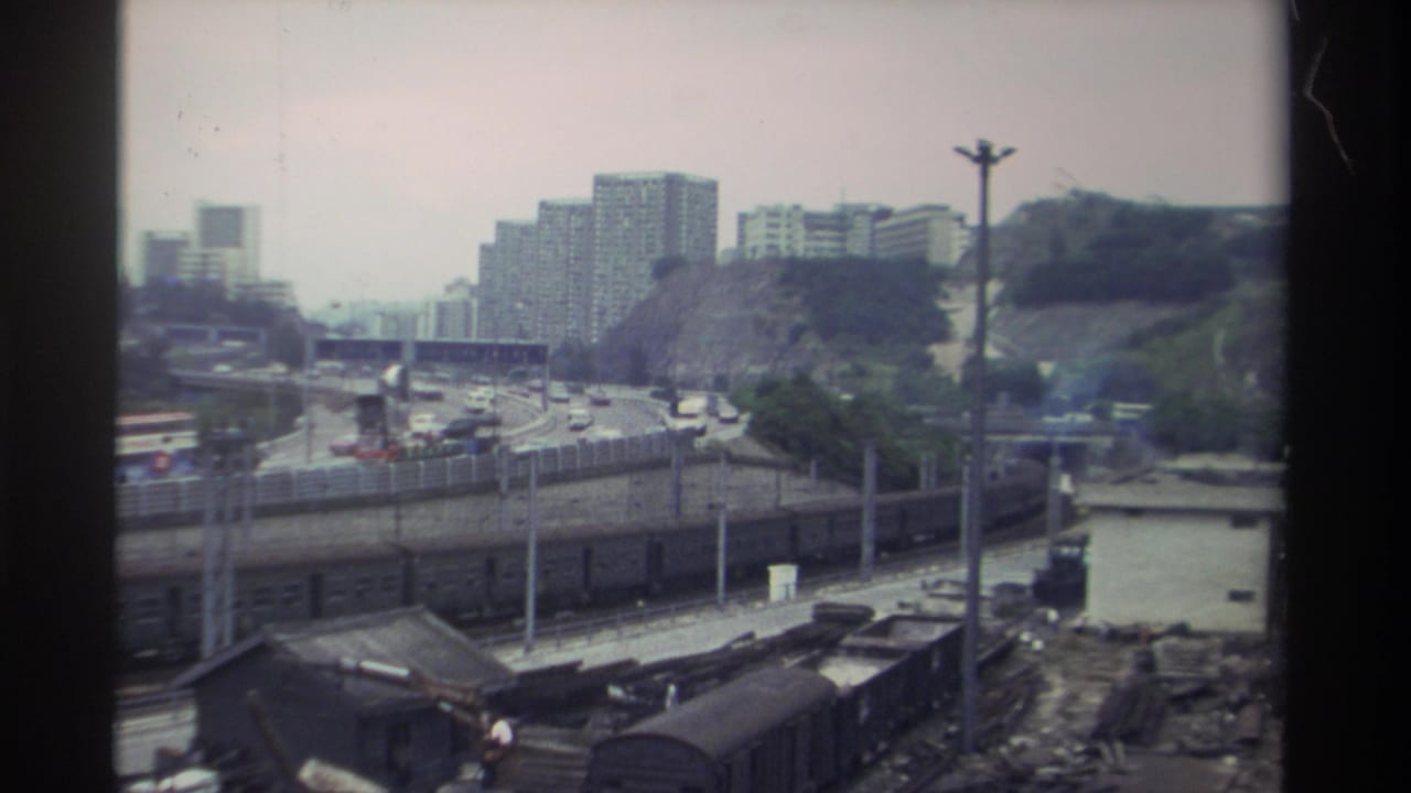 Elevated view of a Taiwan freight rail yard, circa 1982, showing parallel tracks with freight wagons, a multi-lane road, and apartment towers on a hillside under a hazy sky.