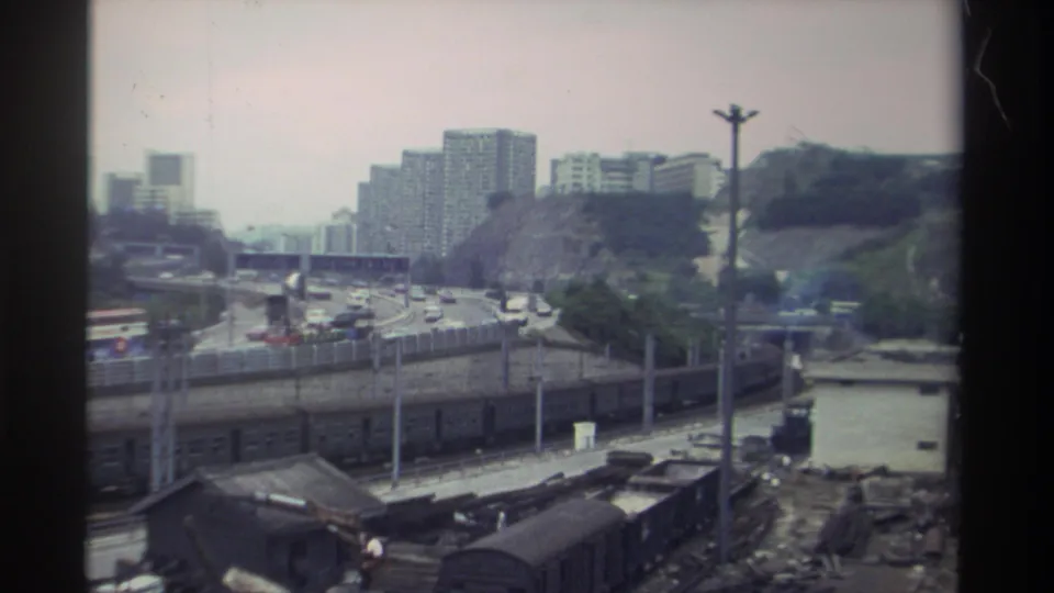 Elevated view of a Taiwan freight rail yard, circa 1982, showing parallel tracks with freight wagons, a multi-lane road, and apartment towers on a hillside under a hazy sky.