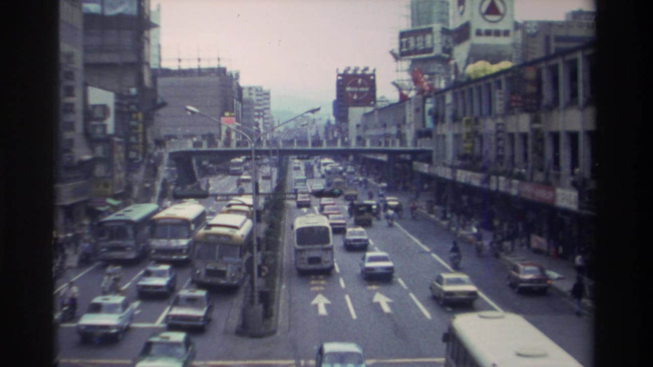 8mm footage looking down a busy Taiwanese urban boulevard circa 1982, with yellow transit buses, mixed traffic, Chinese-character signage, and an elevated walkway spanning the road.