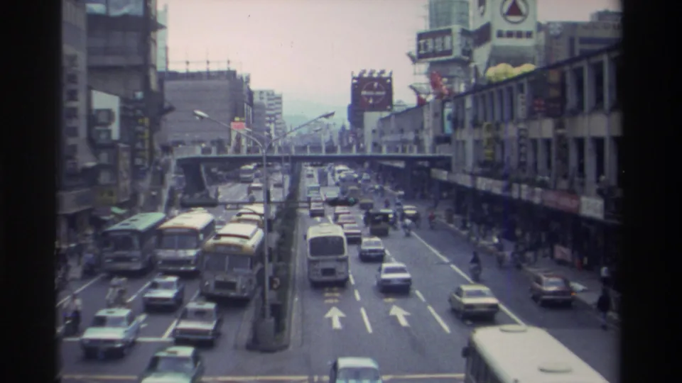 8mm footage looking down a busy Taiwanese urban boulevard circa 1982, with yellow transit buses, mixed traffic, Chinese-character signage, and an elevated walkway spanning the road.