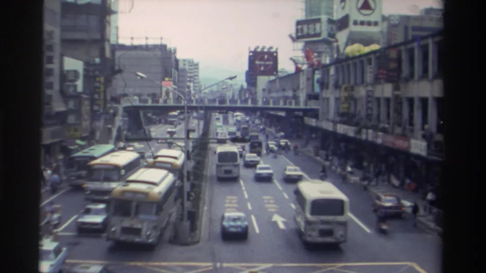8mm frame of a busy Taiwanese city street circa 1982 showing yellow public buses, 1980s sedans, a pedestrian overpass, and Chinese-language storefront signs.