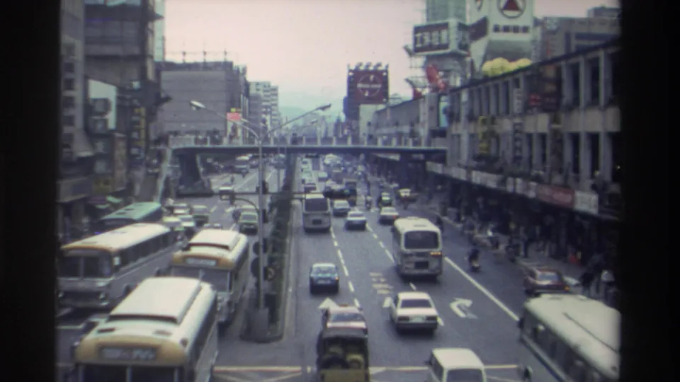 8mm film frame of a multi-lane Taiwanese boulevard circa 1982, with yellow buses in motion, 1980s sedans, Chinese storefronts, and a pedestrian overpass spanning the road.