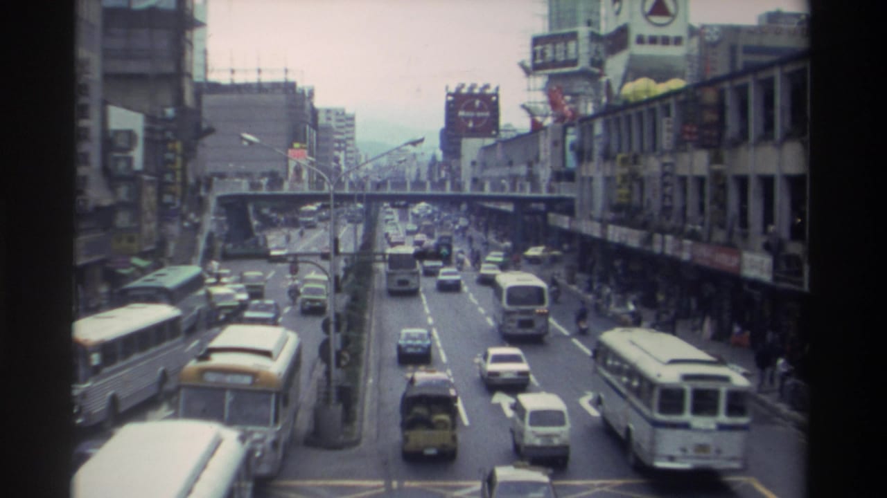Close elevated view of a congested Taiwanese city boulevard in 1982, with white buses and sedans in the foreground and a pedestrian overpass spanning the road mid-distance.