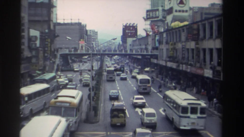 Close elevated view of a congested Taiwanese city boulevard in 1982, with white buses and sedans in the foreground and a pedestrian overpass spanning the road mid-distance.