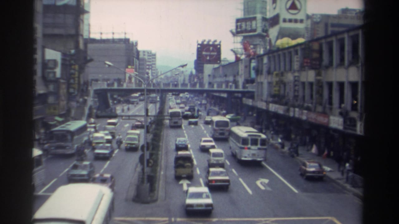 Elevated view along a multi-lane Taiwanese city street with a bus on the right, cars throughout, and a pedestrian overpass visible further along the road.