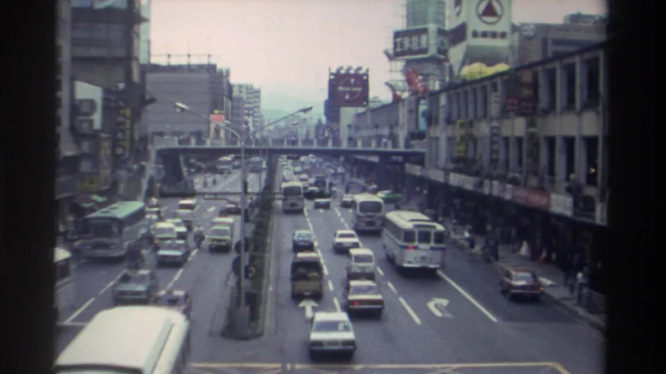Elevated view along a busy Taiwanese urban boulevard in 1982, showing a bus on the nearside lane, mixed car traffic, a pedestrian overpass, and Chinese commercial signage on both sides.