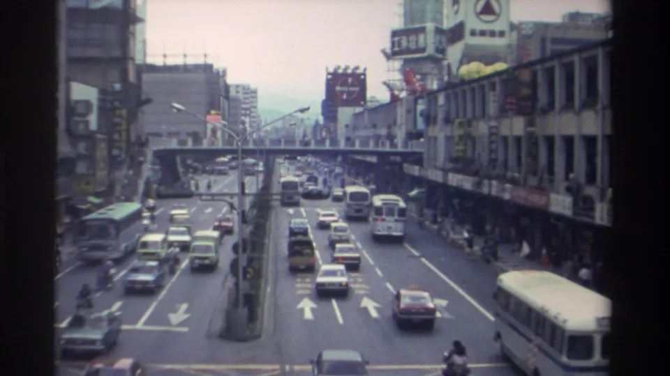 Wider elevated view of a Taiwanese urban boulevard circa 1982, with multi-lane traffic, arrow road markings in the foreground, a pedestrian overpass mid-street, and Chinese-character commercial signage.