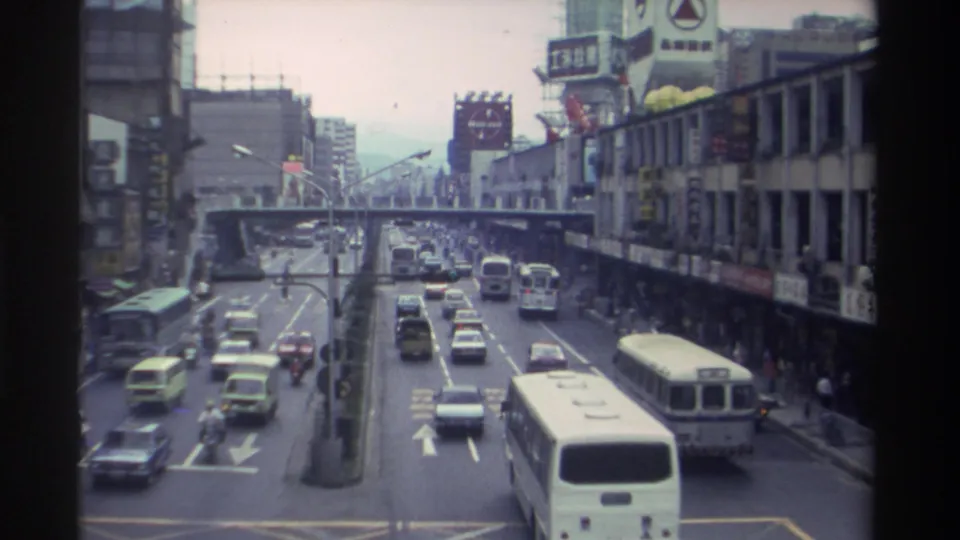 Late-clip elevated view of the same Taiwanese urban boulevard in 1982, with a white bus on the right lane, mixed traffic, and dense Chinese-character commercial signage lining both sides.
