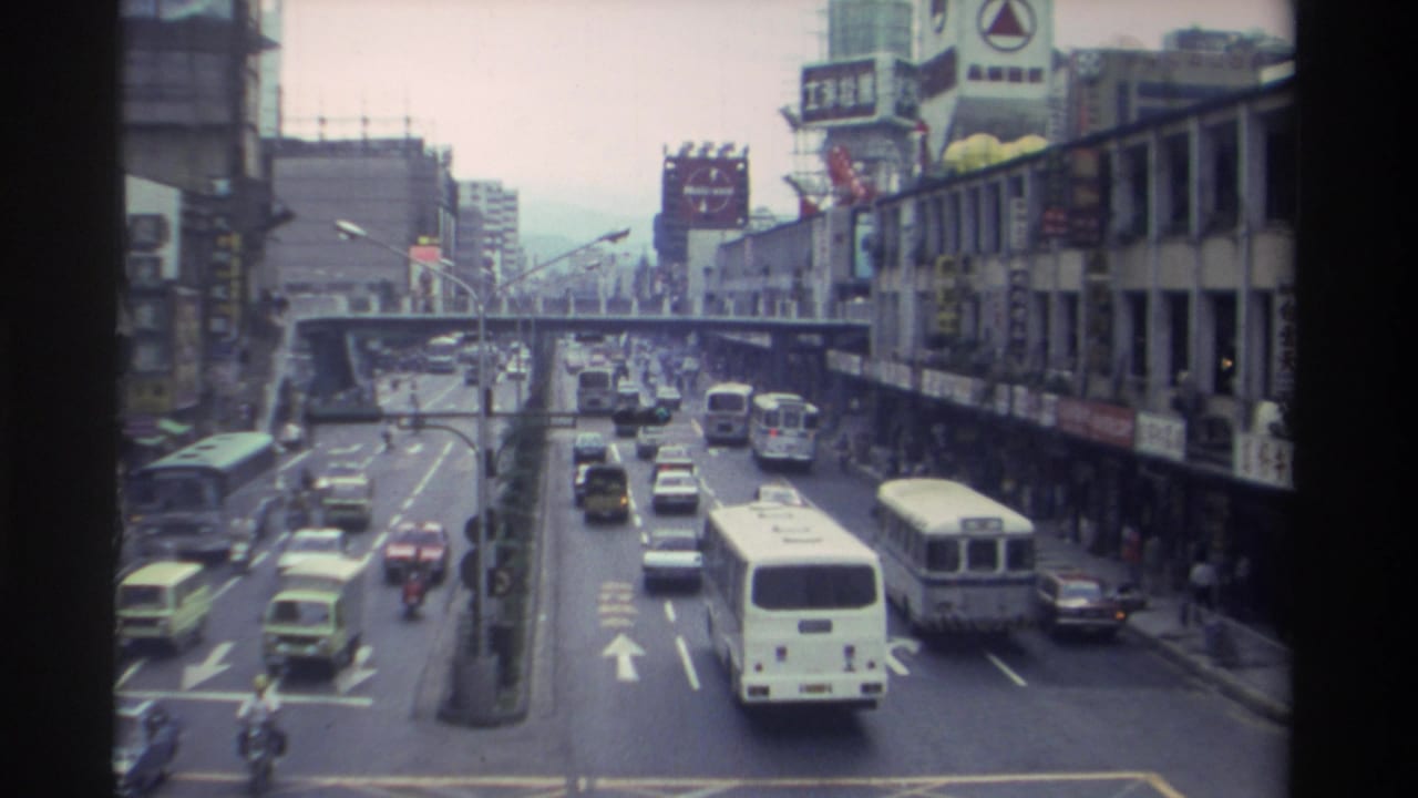 High-angle 8mm shot of a busy Taiwanese city street circa 1982, showing transit buses, sedans, and motorbikes with Chinese-character billboards lining both sides.