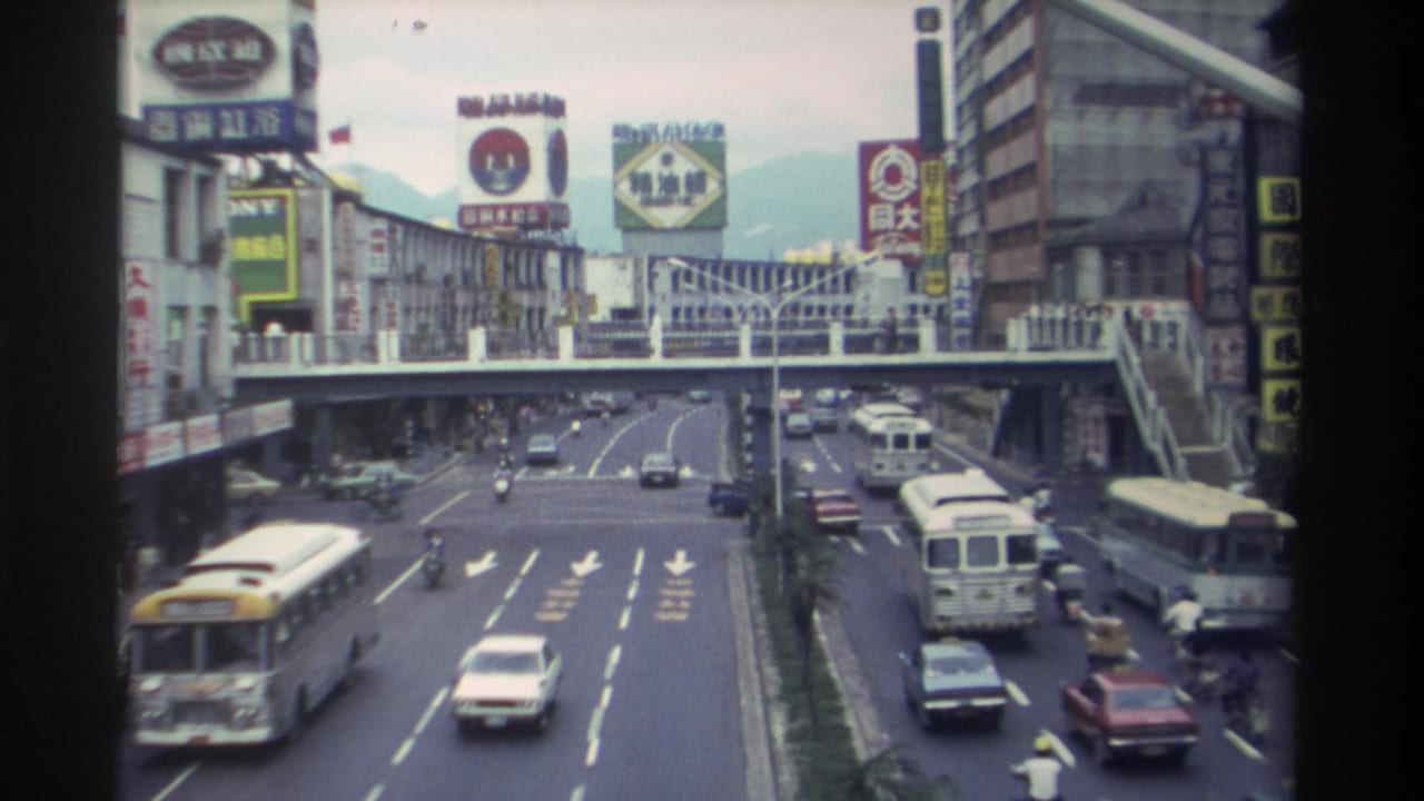 8mm film still from Taiwan circa 1982 showing a multi-lane boulevard with buses, sedans, and motorbikes beneath a pedestrian overpass, with colorful Chinese-character billboard signs and mountains in the background.