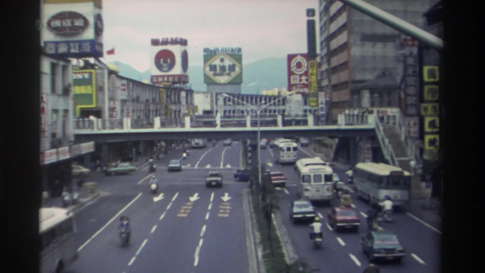 8mm archival footage from Taiwan circa 1982 showing a wide arterial road with mixed traffic beneath a pedestrian overpass, surrounded by dense commercial signage and distant mountains.