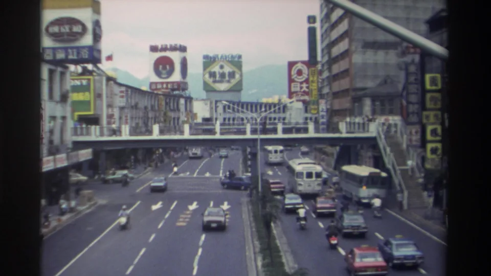 Closing 8mm frame of a Taiwanese city arterial road circa 1982, with buses, sedans, and motorbikes in mixed traffic beneath dense commercial signage; mountains faintly visible in the distance.