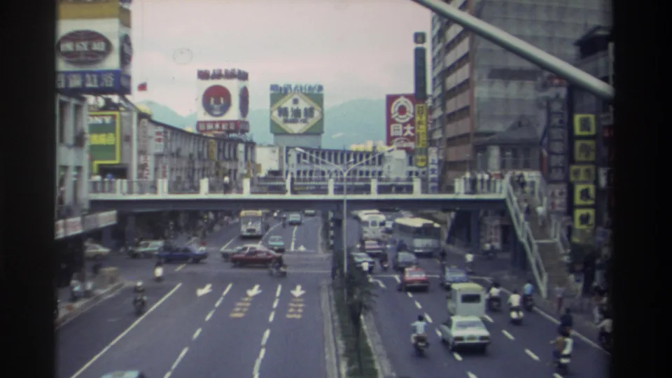 Overhead view of a busy Taiwanese urban boulevard circa 1982, showing city buses, motorcycles, and mid-rise buildings lined with Chinese-character commercial signs.