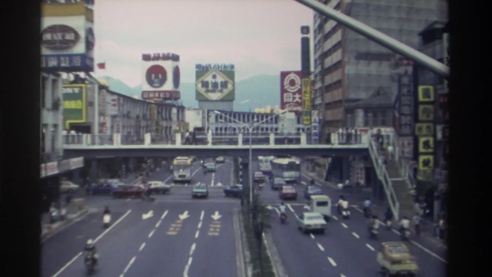 Late-clip frame showing traffic flow on a multi-lane Taiwanese boulevard circa 1982, with scooters prominent among vehicles and a mountain range visible in the distance.