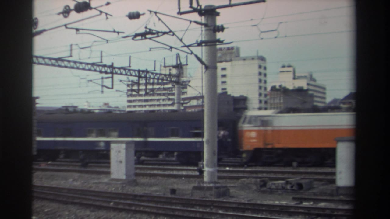 Orange and white locomotive pulling blue carriages past concrete catenary poles, apartment buildings visible in background.
