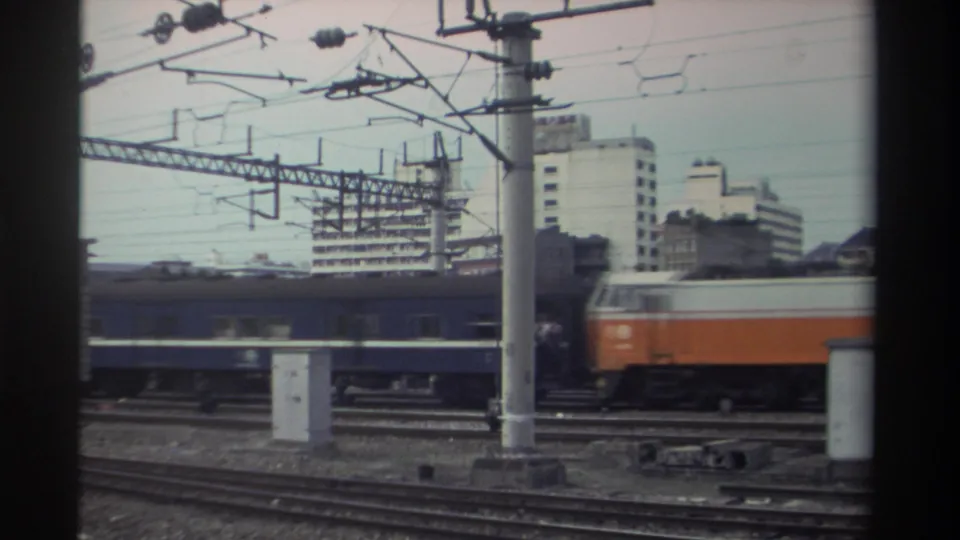 Orange-and-white electric locomotive with blue passenger carriages on electrified urban track, Taiwan, circa 1982; overhead catenary wires and mid-rise buildings behind.