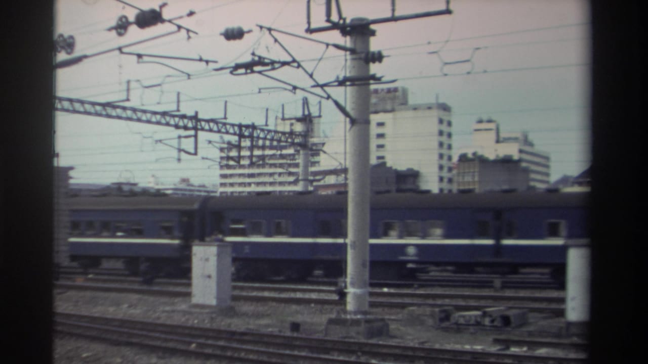 Orange-and-white locomotive with blue passenger carriages continuing through electrified urban rail yard, Taiwan, circa 1982; catenary gantries span the track overhead.
