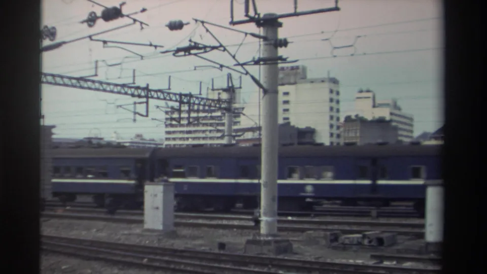 Orange-and-white locomotive with blue passenger carriages continuing through electrified urban rail yard, Taiwan, circa 1982; catenary gantries span the track overhead.