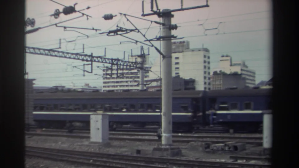 Blue passenger carriages below prominent catenary infrastructure on electrified urban rail corridor, Taiwan, circa 1982; apartment blocks visible beyond the tracks.