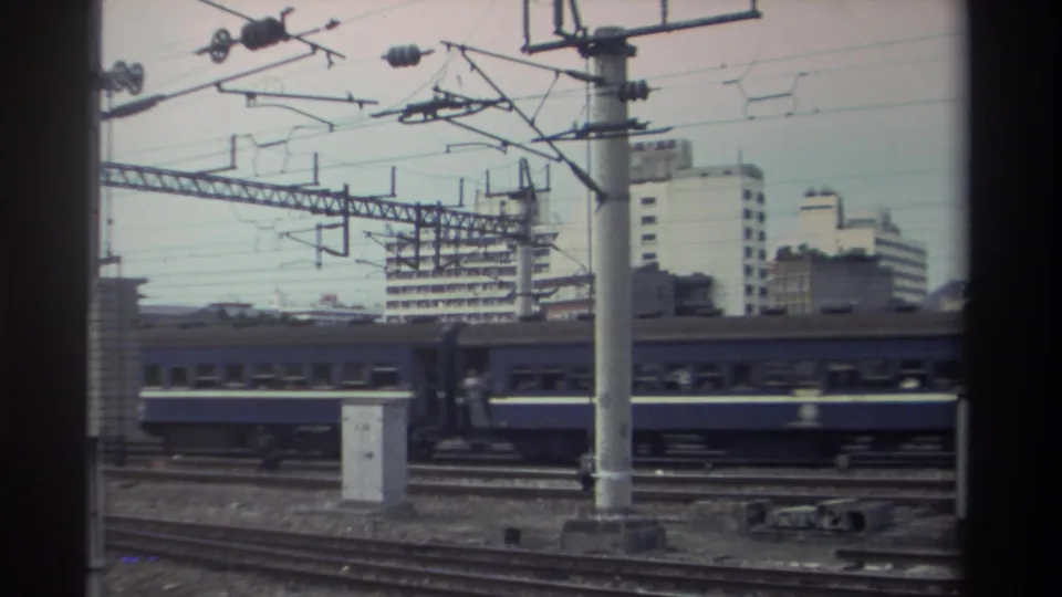 Electrified rail yard with blue passenger carriages, concrete catenary poles and wire gantry arms, mid-rise buildings beyond, Taiwan, circa 1982.