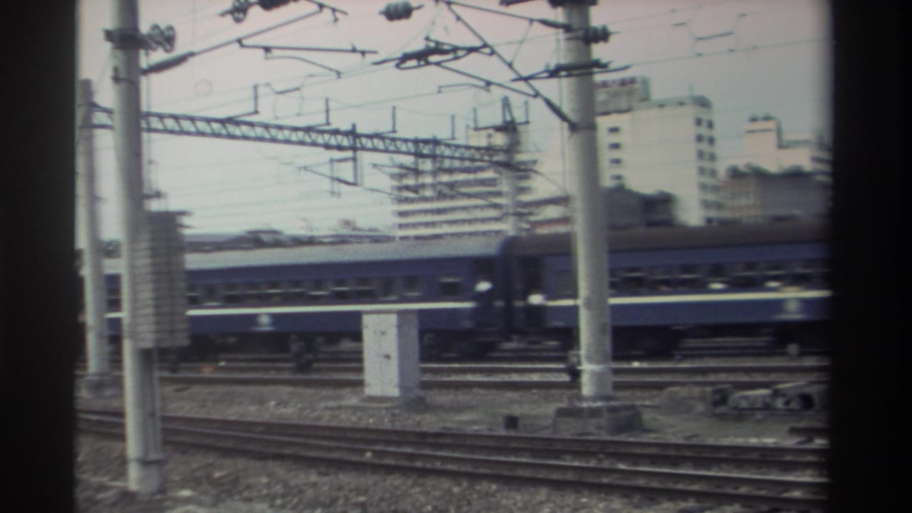 Blue passenger train passing through electrified rail yard in urban Taiwan, circa 1982; rear coaches visible at right as train exits frame.