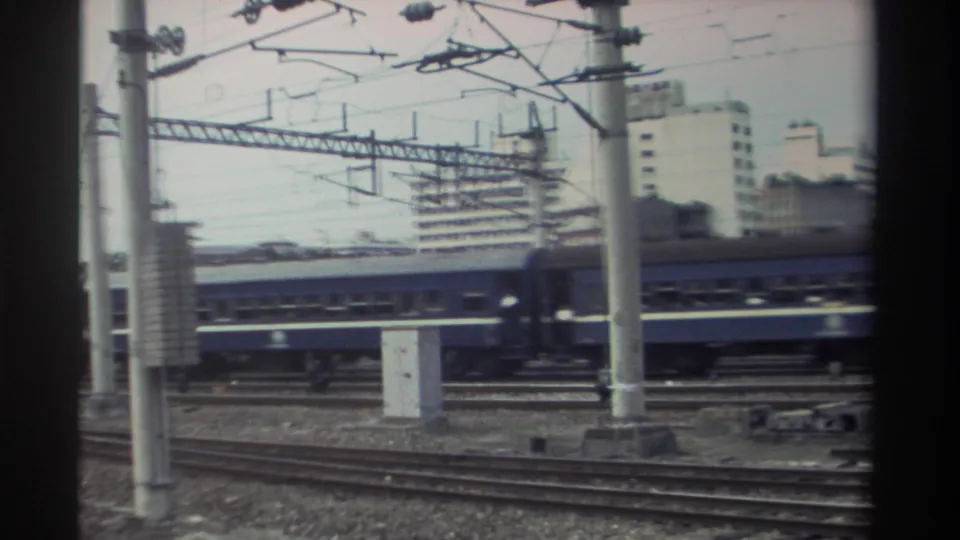 Blue passenger train passing through electrified rail yard in urban Taiwan, circa 1982; rear coaches visible at right as train exits frame.