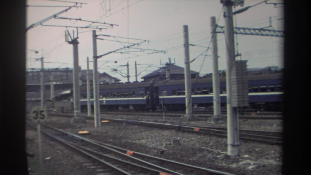 Elevated view of a rail yard with multiple parallel tracks, a blue passenger train, and overhead electric catenary lines on poles.