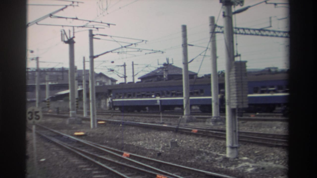 Elevated rail yard view with parallel tracks, blue passenger train, and overhead catenary lines; nearly identical angle to preceding frame.