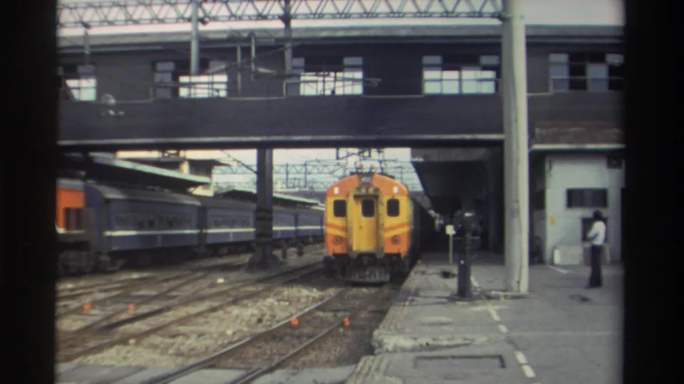 Taiwan railway station platform, circa 1982; an orange and yellow locomotive pulls under a covered canopy while a lone figure stands on the platform below.