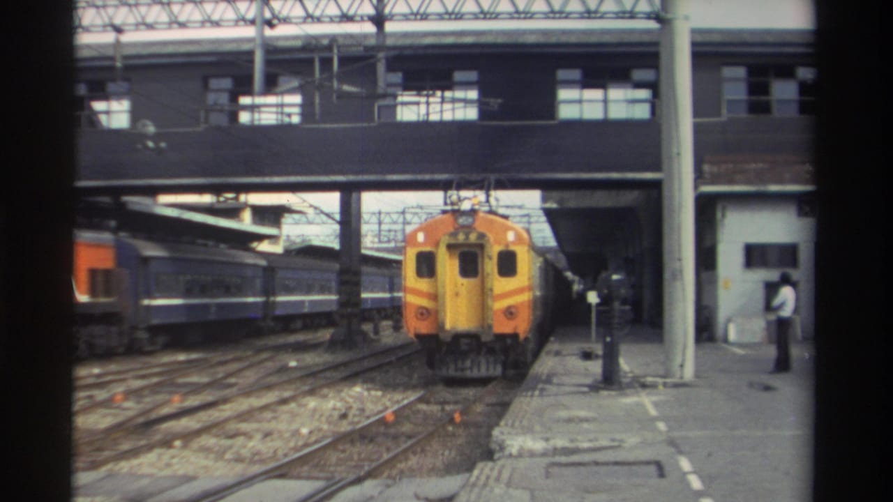 Orange and yellow train arrives at a Taiwanese railway station, 1982; concrete platform, overhead bridge, and standing figure visible on 8mm film.