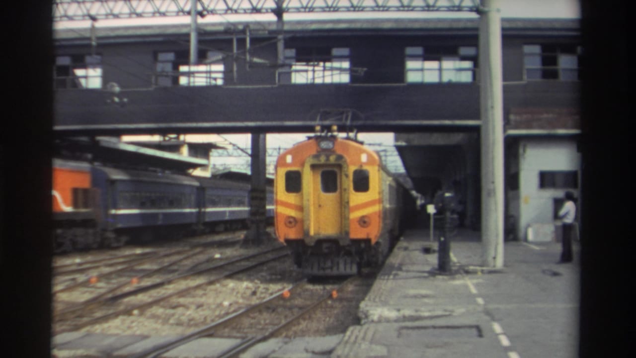 Orange and yellow train advancing toward camera; figure in light clothing stands on right platform.