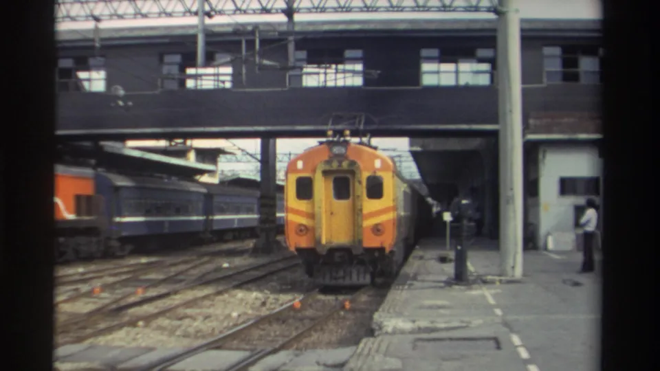Orange and yellow train advances through a Taiwanese station, 1982; a standing figure is visible on the right platform.