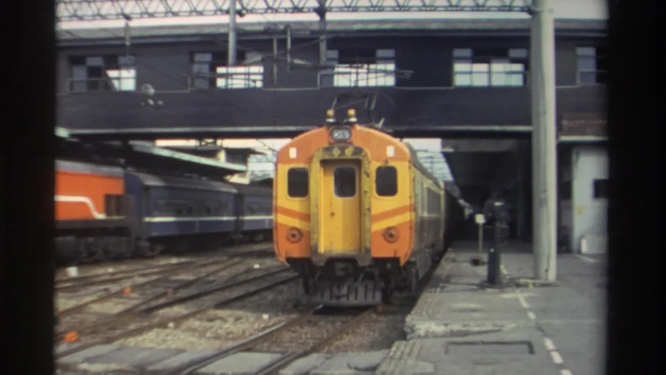Close approach of orange and yellow train at a Taiwanese station, 1982; platform pillars and overhead structure frame the shot.