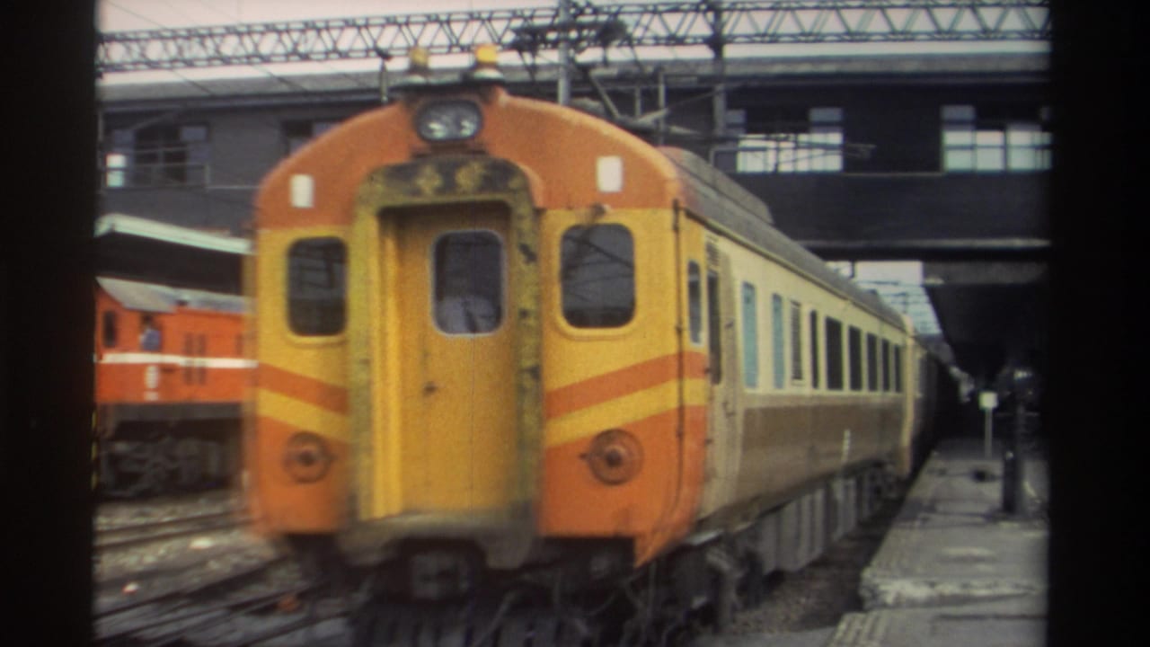 Orange-and-yellow diesel locomotive at a covered Taiwan railway station, circa 1982, with cream-and-brown passenger coaches trailing behind, filmed on 8mm.