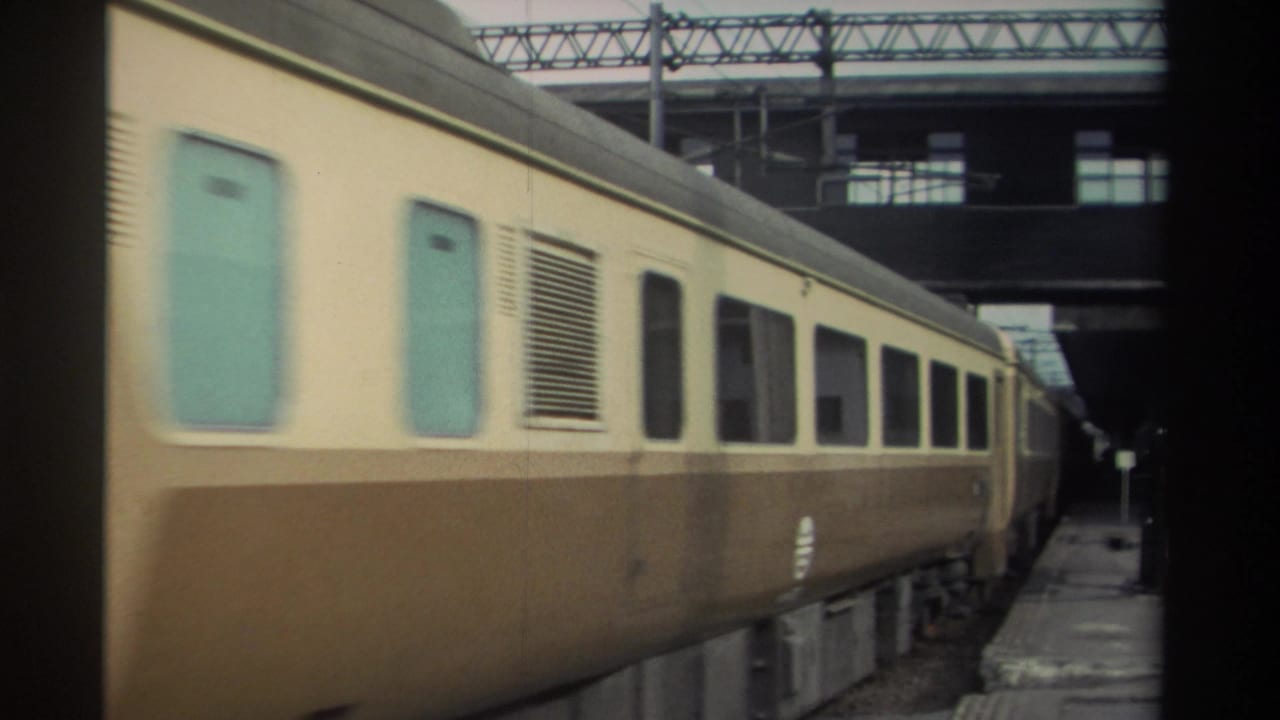 Side view of cream-and-brown passenger coaches passing at speed, turquoise door panels visible, overhead electrification gantry above.