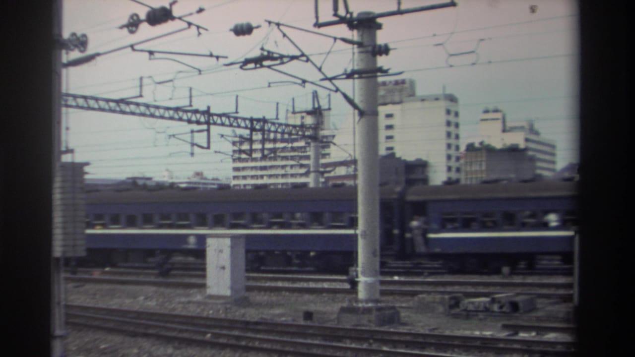 Blue passenger train moving right on electrified tracks; large catenary pole in foreground, mid-rise buildings beyond.