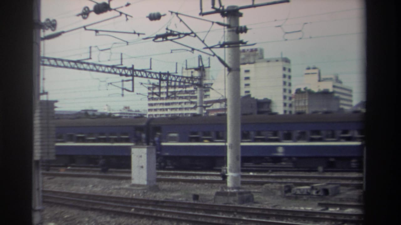 Blue passenger train continuing past electrified rail infrastructure; catenary pole centered, buildings in background.