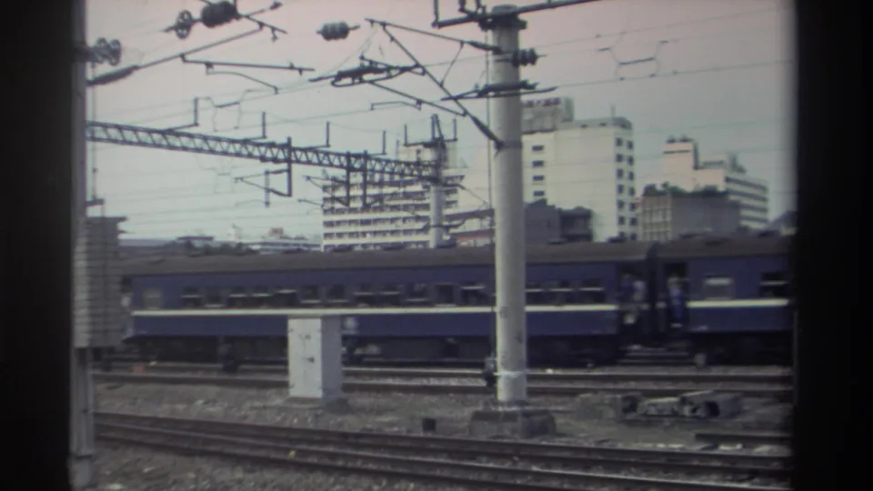 Blue passenger train on electrified tracks, Taiwan, c.1982; catenary pole foreground, prominent white mid-rise building and rail yard visible.