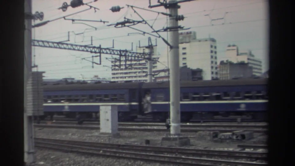 Blue passenger train at end of frame pass, Taiwan, c.1982; catenary pole and wires prominent, mid-rise concrete buildings and rail infrastructure visible.