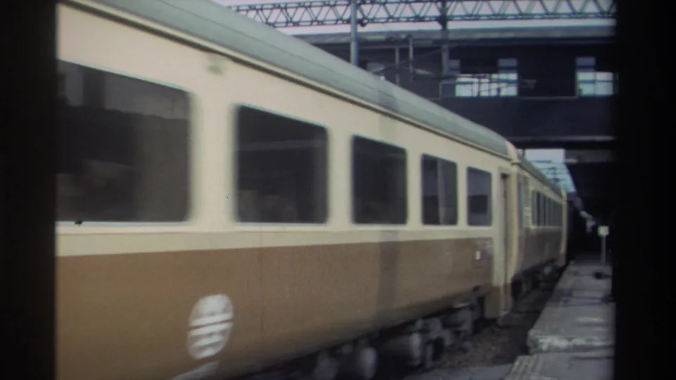 Cream-and-brown passenger train cars beneath a metal lattice canopy at a station platform, Taiwan, 1982, 8mm.