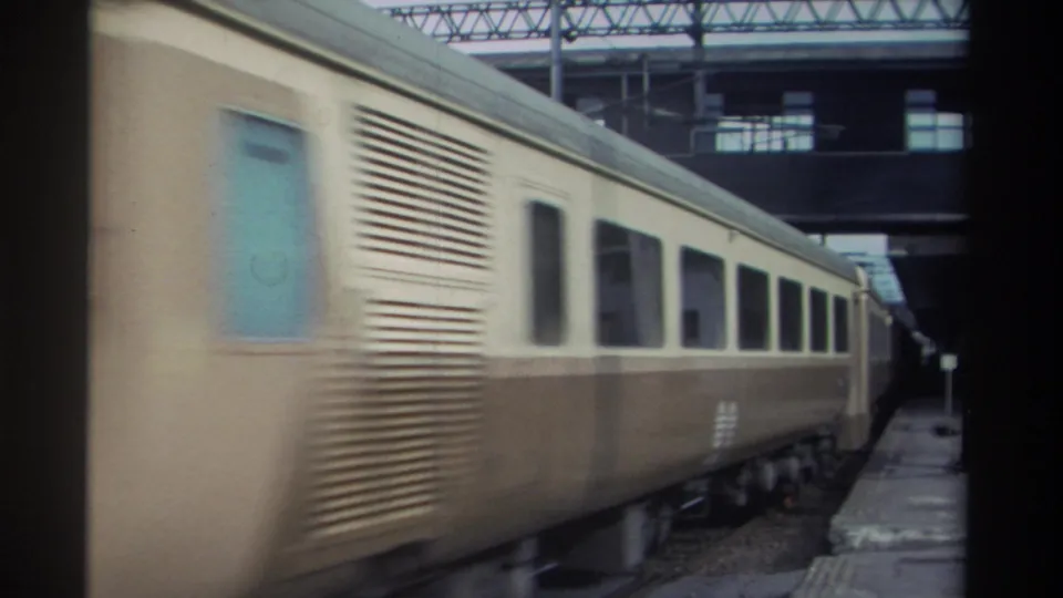 Side of a passenger train car with louvered panels and blue door passing at speed under a station canopy, Taiwan, 1982, 8mm.