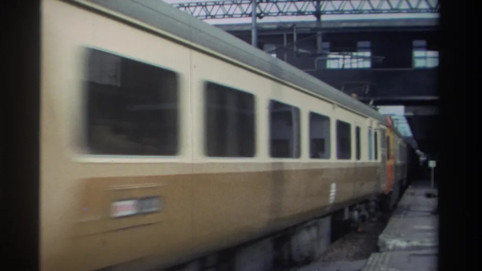 Cream-and-brown passenger cars moving past a station platform in Taiwan, circa 1982, with overhead rail infrastructure and a second train visible beyond.