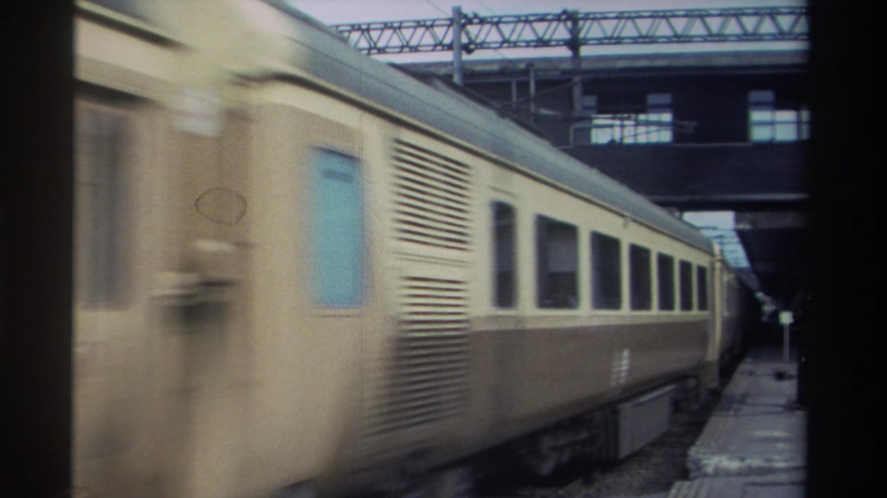 Cream and brown passenger train moving along a covered station platform, shot handheld from platform level with motion blur at the leading cars.