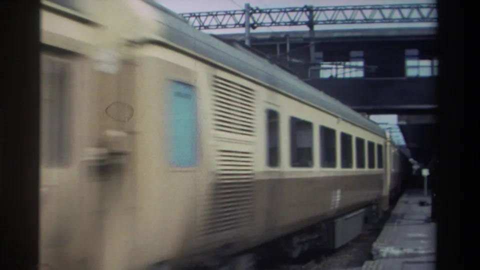 Handheld 8mm shot of a two-tone cream-and-brown passenger train passing a covered station platform in Taiwan, circa 1982, overhead steel truss structure visible.