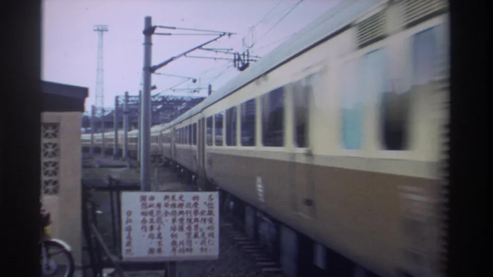 Side of a cream-and-brown passenger train at a Taiwan station, 1982, with overhead catenary infrastructure, a Chinese-script signage board, and a parked bicycle visible.