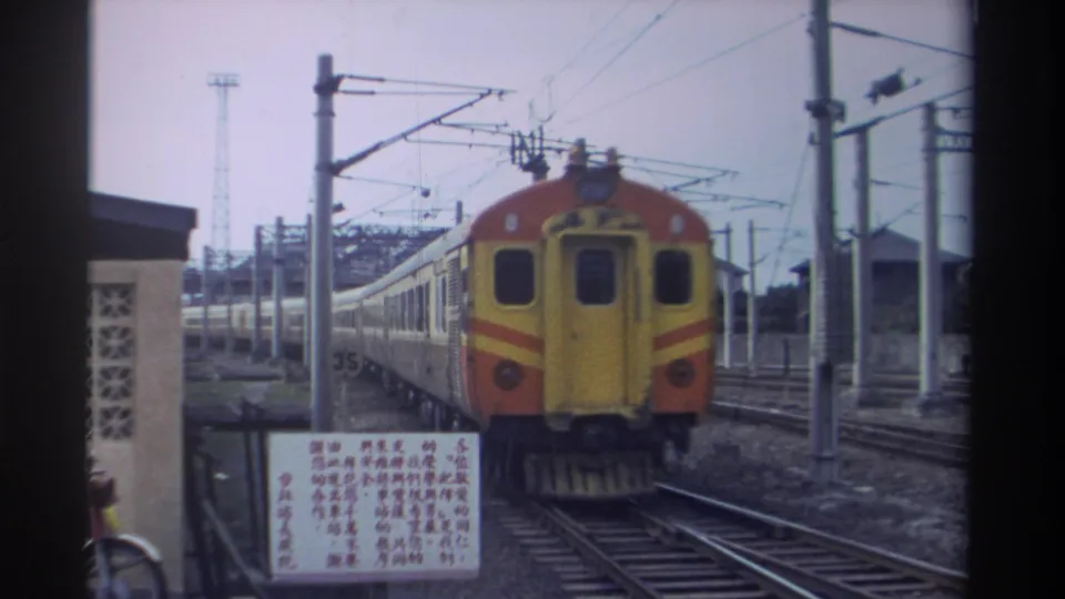 Yellow-and-orange locomotive approaching camera at a Taiwan station, 1982, with catenary poles and wires, Chinese-script platform signage, multiple tracks, and a lattice-block station building.