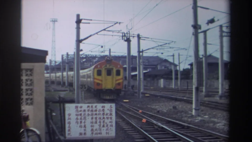 Yellow-and-orange locomotive at a Taiwan station, 1982, viewed from the platform with overhead catenary infrastructure, Chinese-script signage, and a lattice-block station building.